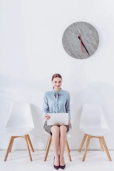 Mujer alegre sentada en la silla con el ordenador portátil mientras espera entrevista de trabajo - foto de stock