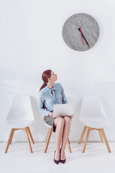 Mitarbeiter sitzt mit Laptop auf Stuhl und schaut auf Uhr — Stockfoto
