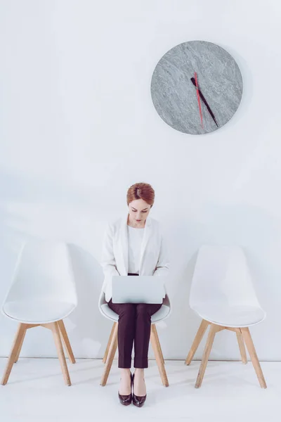 Donna attraente utilizzando il computer portatile mentre seduto sulla sedia vicino orologio — Foto stock