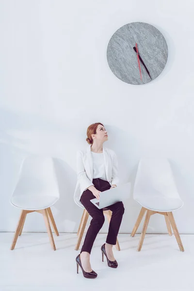 Beautiful employee sitting on chair with laptop and looking at clock — Stock Photo