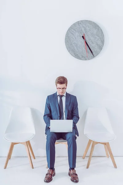 Bell'uomo in occhiali seduto sulla sedia e utilizzando il computer portatile vicino all'orologio — Foto stock