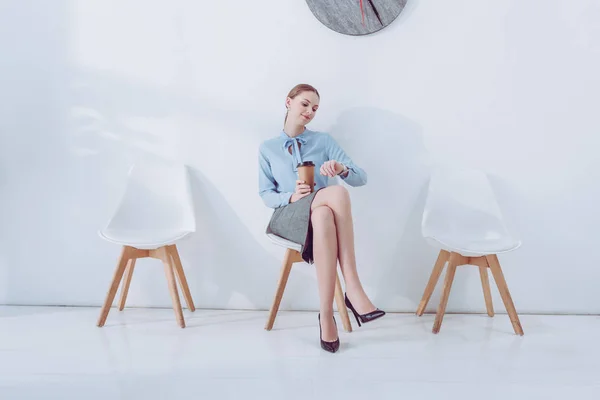 Attraente dipendente in possesso di tazza di carta e guardando orologio mentre seduto sulla sedia — Foto stock