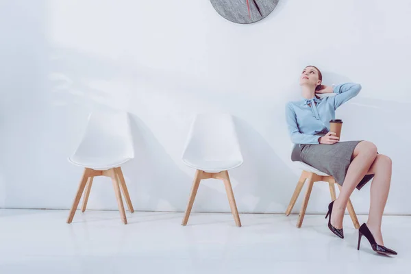 Приваблива жінка тримає паперову чашку і дивиться на годинник, сидячи на стільці — стокове фото