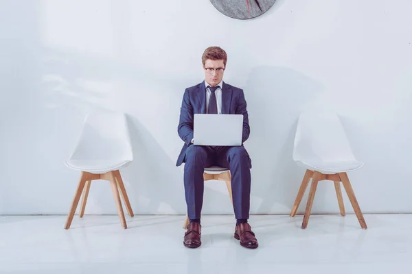 Empleado guapo en gafas sentado en la silla y el uso de la computadora portátil - foto de stock