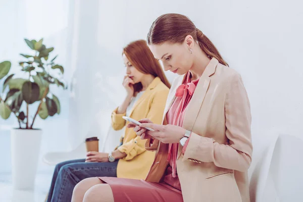 Селективный фокус красивой женщины с помощью смартфона рядом с сотрудником с бумажной чашкой — стоковое фото