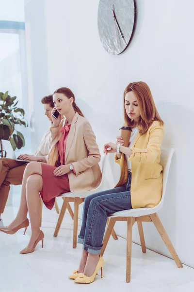 Mitarbeiter sitzt mit Pappbecher und schaut auf Uhr in der Nähe von Mann und Frau mit Gadgets — Stockfoto