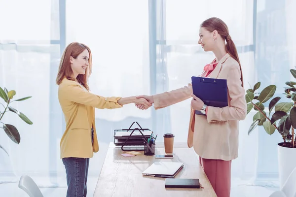 Happy recruiter holding clipboard and shaking hands with woman in modern office — Stock Photo
