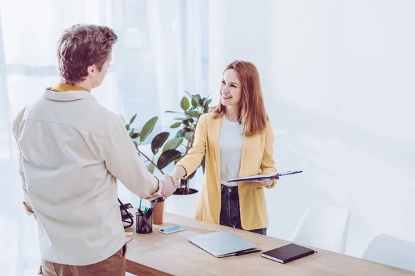 Happy recruiter holding clipboard and shaking hands with employee in modern office — Stock Photo