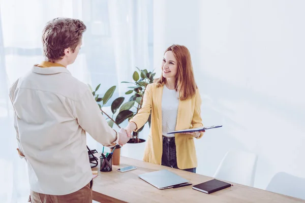 Recrutador feliz segurando prancheta e apertando as mãos com o empregado no escritório moderno — Fotografia de Stock