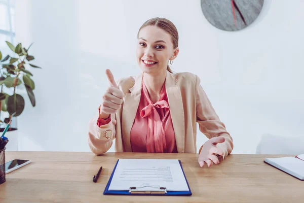 Fröhlicher Recruiter zeigt Daumen hoch, während er in der Nähe des Klemmbretts sitzt und in die Kamera schaut — Stockfoto