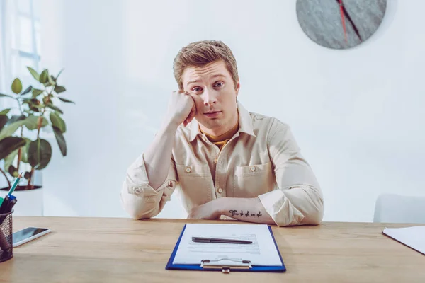 Gelangweilt gut aussehender Personalvermittler sitzt neben Klemmbrett und Stift und schaut in die Kamera — Stockfoto