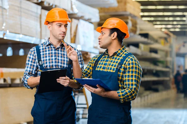 Dos trabajadores de almacenes multiculturales serios hablando y sosteniendo portapapeles - foto de stock