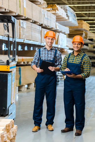 Two smiling multicultural workers in overalls and safety vasts smiling and looking at camera — Stock Photo