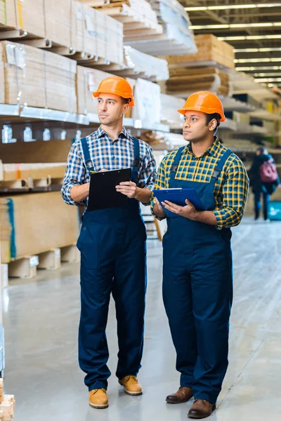 Dos atentos trabajadores de almacenes multiculturales en escritura uniforme en portapapeles - foto de stock