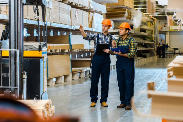 Serious storehouse worker in uniform pointing with finger while standing near indian colleague — Stock Photo