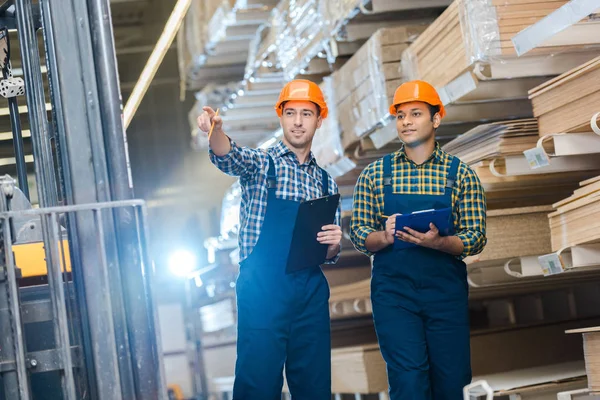 Dos trabajadores multiculturales en camisas a cuadros y botes de seguridad con portapapeles - foto de stock