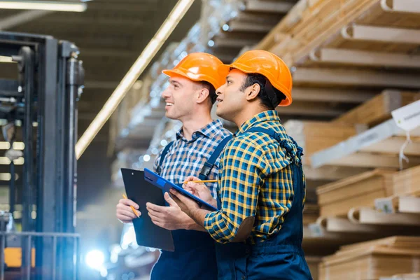 Dos trabajadores de almacenes multiculturales sonriendo mientras sostienen portapapeles - foto de stock