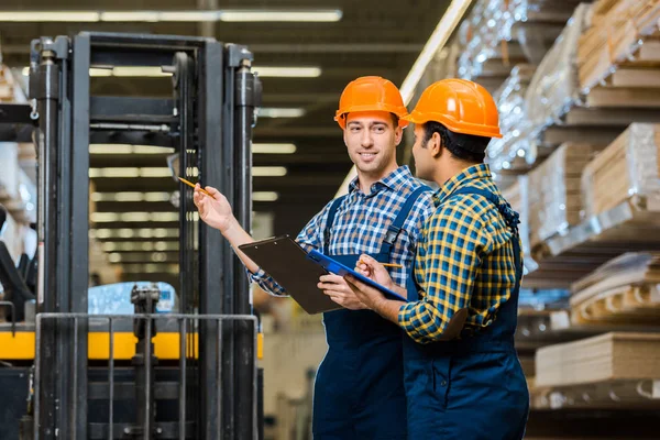 Smiling warehouse worker in uniform and helmet talking to indian colleague — Stock Photo