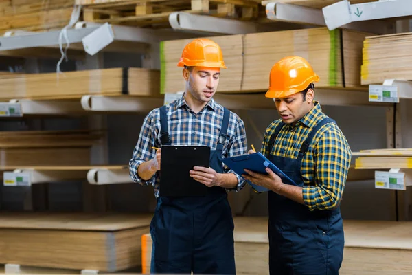 Dos trabajadores de almacenes multiculturales en escritura uniforme en portapapeles - foto de stock