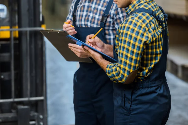 Partial view of two multicultural warehouse workers in plaid shirts writing in clipboards — Stock Photo