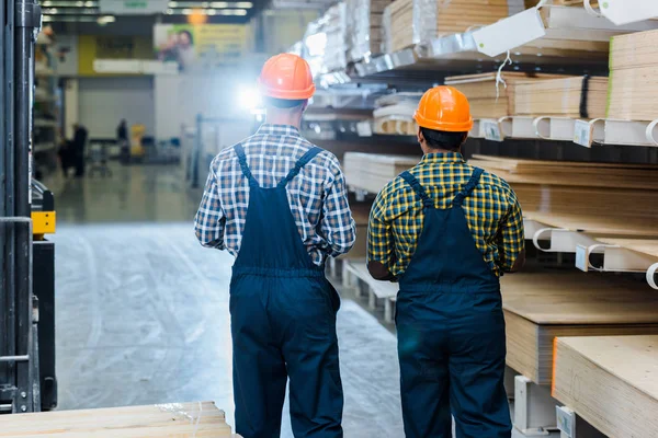 Visão traseira de dois trabalhadores multiculturais em macacões, camisas xadrez e capacetes no armazém — Fotografia de Stock