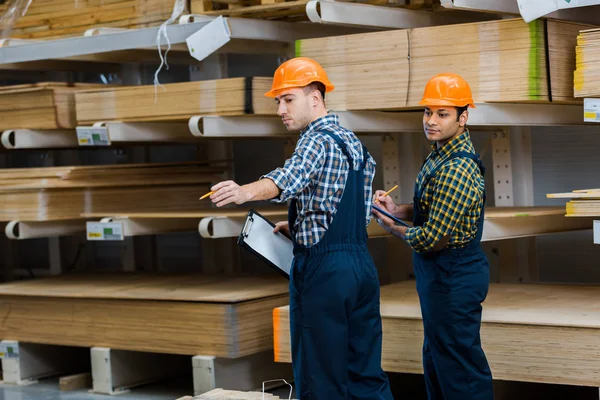 Deux travailleurs multiculturels en uniforme debout près de matériaux de construction en bois — Photo de stock