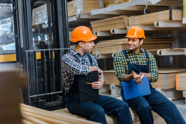 Enfoque selectivo de dos trabajadores multiculturales sonrientes sentados en el almacén - foto de stock