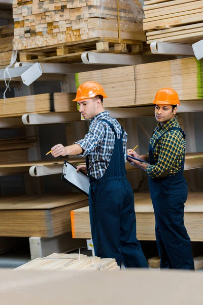 Dos trabajadores multiculturales con portapapeles de pie cerca de materiales de construcción de madera - foto de stock