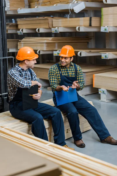 Zwei lächelnde multikulturelle Arbeiter mit Klemmbrettern im Gespräch, während sie im Lager sitzen — Stockfoto