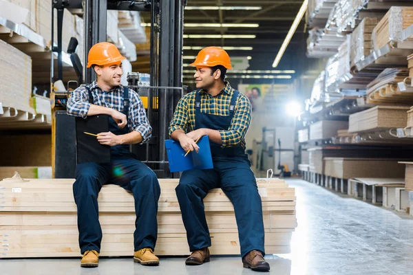 Two multicultural workers smiling and talking while sitting on plywood in warehouse — Stock Photo