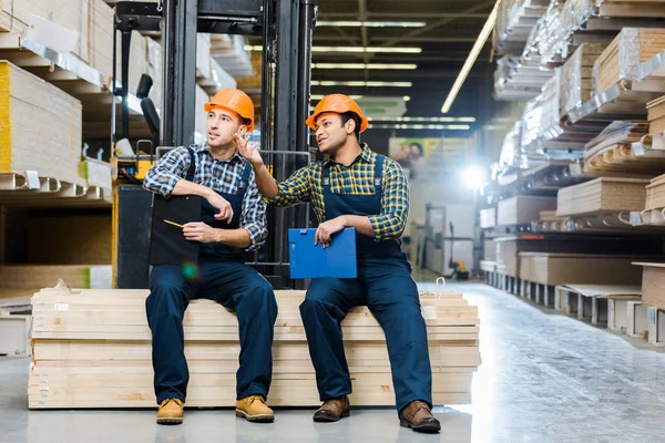 Two multicultural workers talking while sitting on plywood in warehouse — Stock Photo