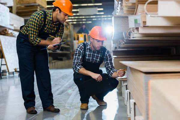 Dos colegas multiculturales en uniforme y cascos trabajando en almacén - foto de stock