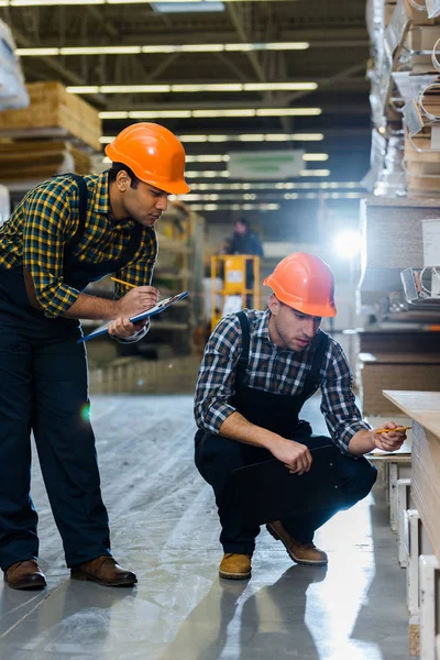 Two multicultural colleagues in uniform and helmets working in warehouse — Stock Photo