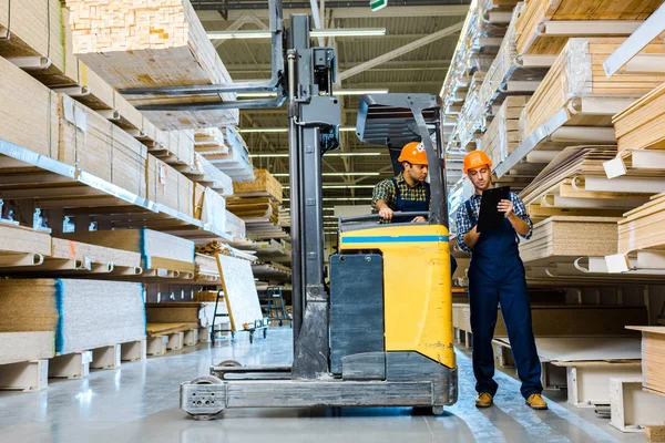 Lagerarbeiter schreibt in Klemmbrett neben indischem Kollegen in Gabelstapler-Maschine — Stockfoto