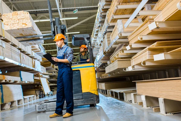 Concentrated indian warehouse worker sitting in forklift machine near thoughtful colleague writing in clipboard — Stock Photo