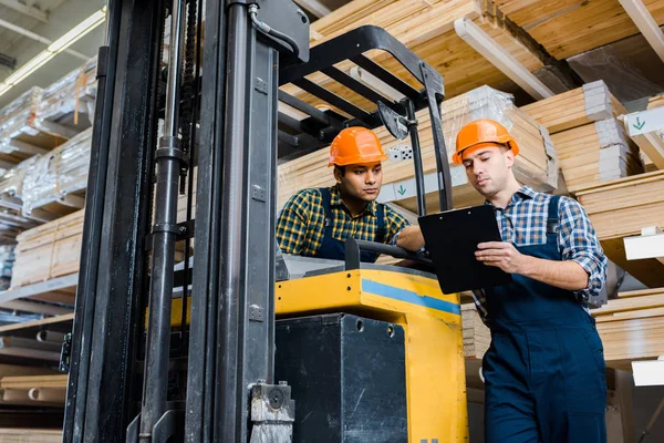 Multicultural warehouse workers talking near forklift machine — Stock Photo