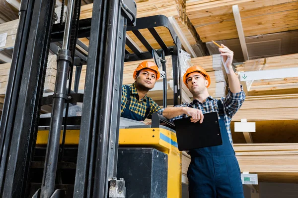 Warehouse worker pointing with pencil near indian colleague sitting in forklift machine — Stock Photo