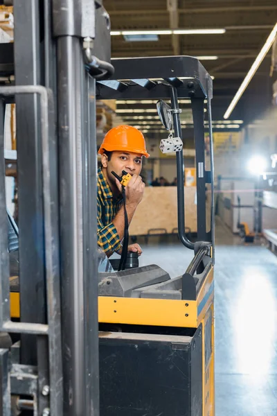 Good-looking indian warehouse worker sitting in forklift machine and talking on walkie talkie — Stock Photo