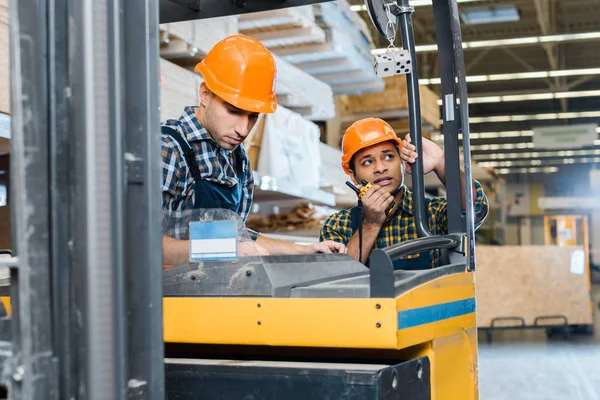 Warehouse worker in helmet sitting in forklift machine near indian colleague talking on walkie talkie — Stock Photo