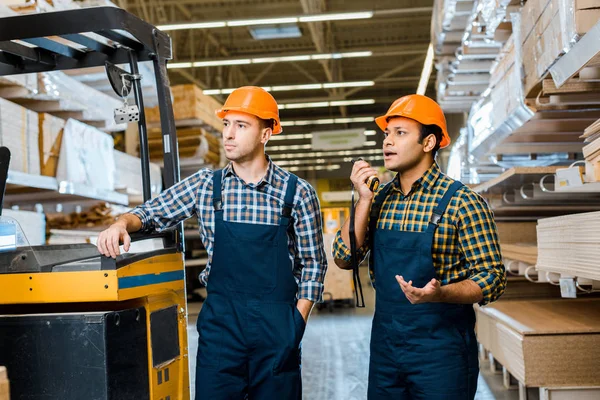 Multicultural warehouse workers in uniform and helmets standing near forklift machine in storehouse — Stock Photo