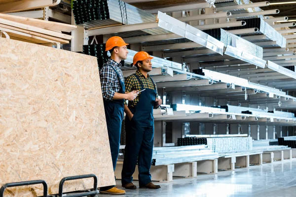 Dos amigos multiculturales en uniforme de pie cerca de bastidores con materiales de construcción metálicos - foto de stock