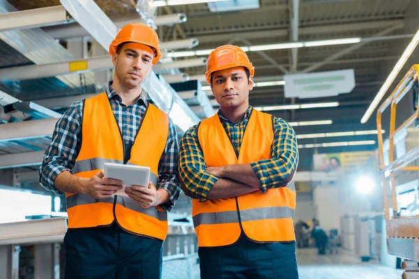 Serious multicultural workers in safety vasts and helmets looking at camera — Stock Photo