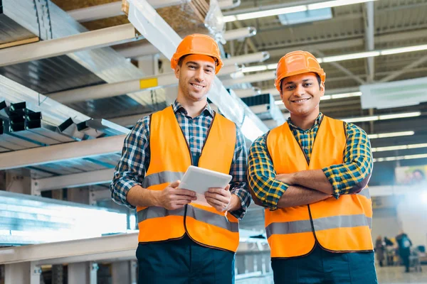 Handsome multicultural workers in safety vasts and helmets smiling and looking at camera — Stock Photo