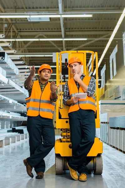 Handsome indian worker pointing with hands while standing with colleague near scissor lift — Stock Photo