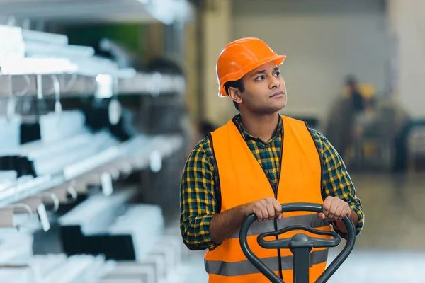 Handsome indian warehouse worker standing near pallet jack and looking away — Stock Photo