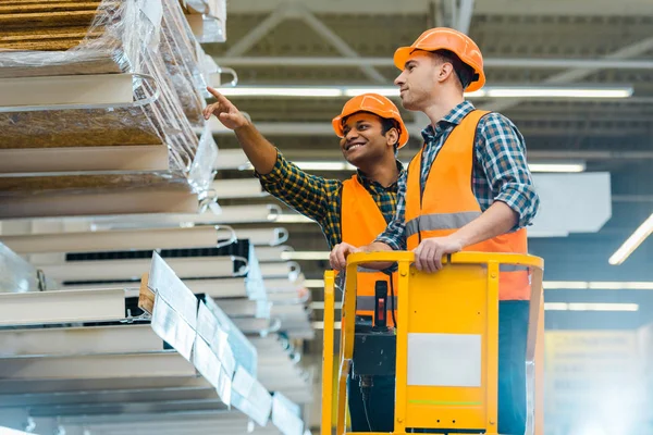 Smiling indian warehouse worker pointing with finger at construction materials while standing on scissor lift near colleague — Stock Photo