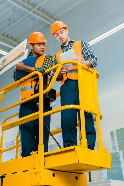 Concentrated multicultural workers with digital tablet and clipboard standing on scissor lift — Stock Photo