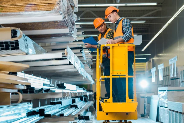 Multicultural workers with digital tablet and clipboard standing on scissor lift near shelves with metallic construction materials — Stock Photo