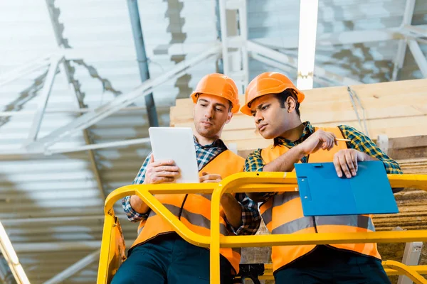 Nachdenkliche Multikulti-Arbeiter nutzen digitales Tablet, während sie im Lager auf einem Scherenaufzug stehen — Stockfoto