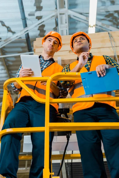 Trabajadores de almacenes multiculturales concentrados con tableta digital y portapapeles de pie sobre tijera elevadora - foto de stock